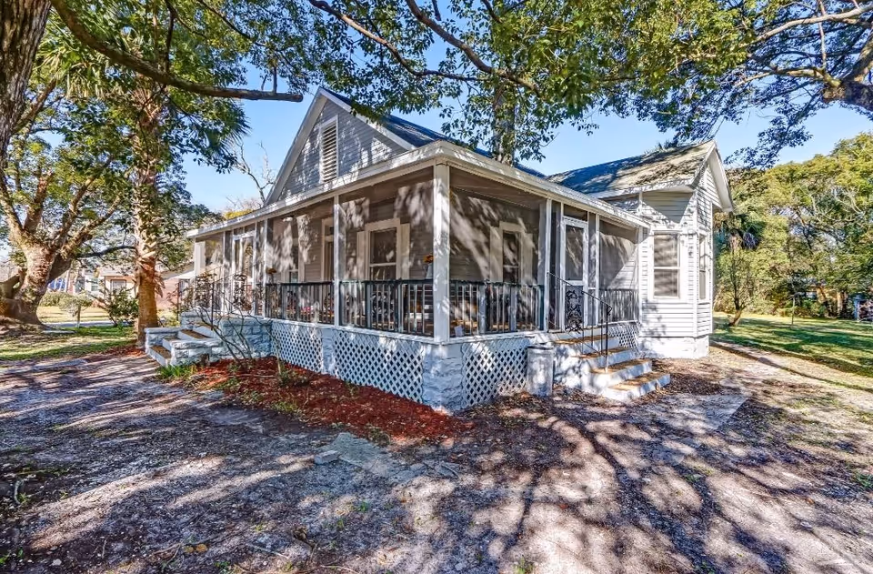 Exterior view of a single-story house with a screened-in porch, surrounded by trees and greenery under a clear blue sky.