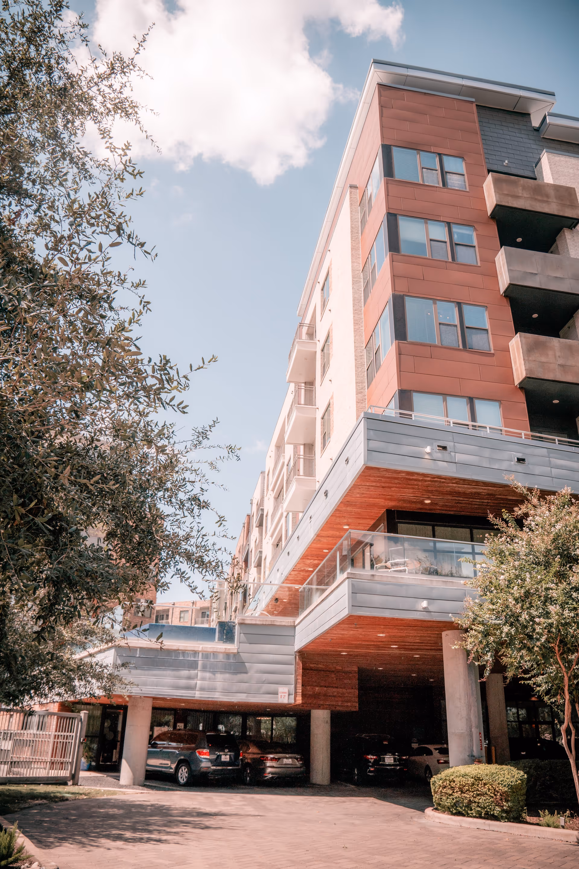 Exterior view of a multi-story residential building with balconies and a covered parking area underneath. Trees and shrubs surround the building, and several cars are parked in the lot. The sky is partly cloudy with blue patches visible.