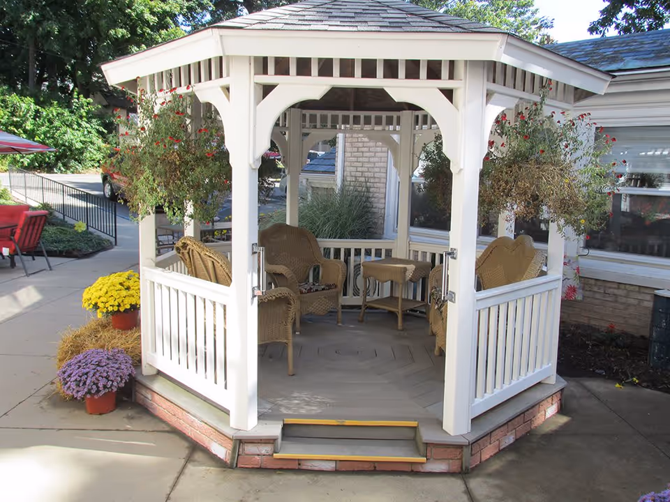 A white wooden gazebo with a shingled roof, containing wicker chairs and a small table inside. The gazebo is surrounded by potted flowers and hanging plants, situated on a concrete patio near a building with windows.