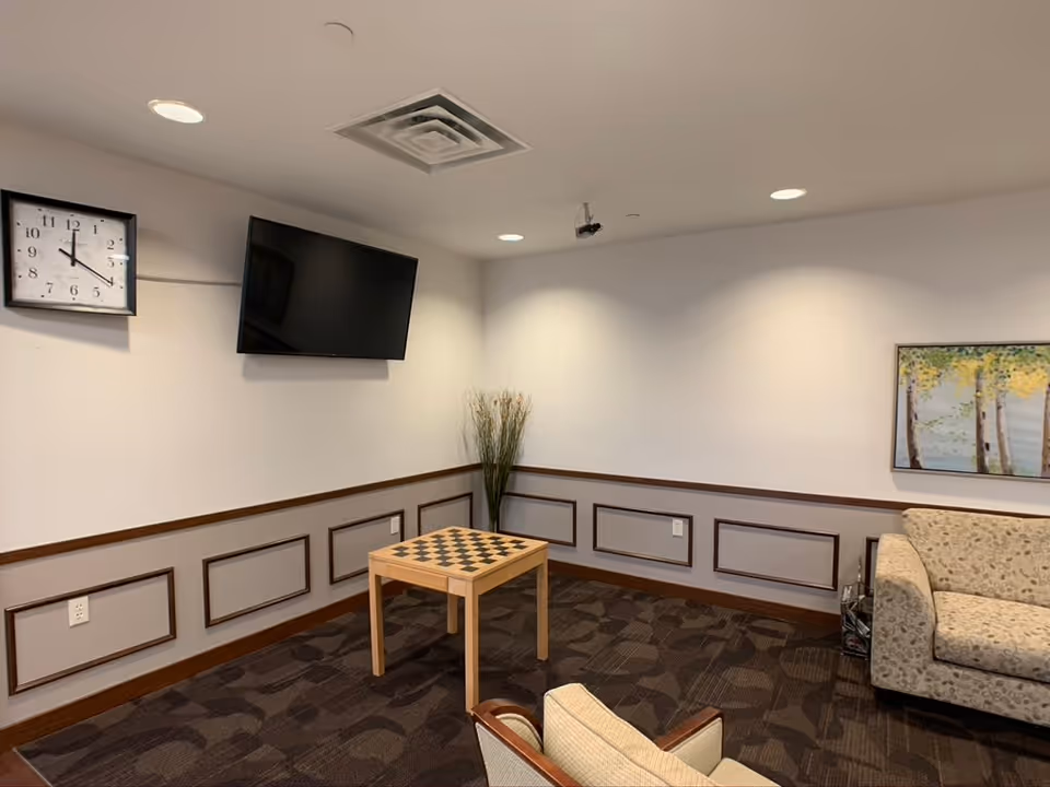 A cozy living room area with a wall-mounted flat screen TV, a clock showing 12:15, a small wooden table with a checkerboard pattern, a beige patterned sofa, a beige armchair, a tall vase with decorative grass, and a framed painting of trees on the wall.