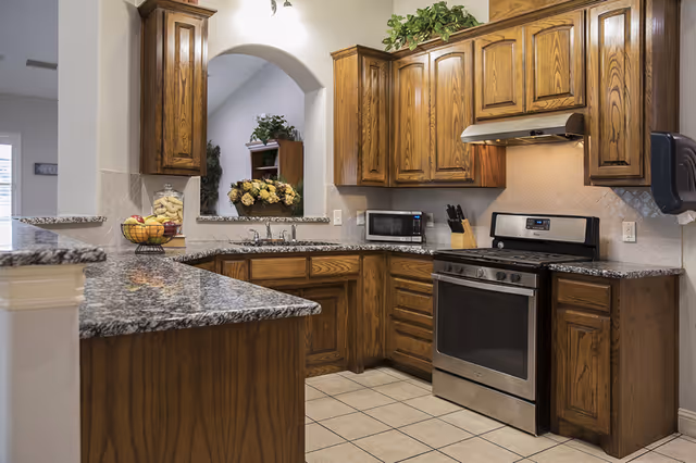 A kitchen with wooden cabinets, granite countertops, a stainless steel gas stove with an overhead vent, a microwave, a knife block, and a fruit basket on the counter. There is a tiled floor and a small arched opening in the wall showing a glimpse of a room with flowers and a plant.