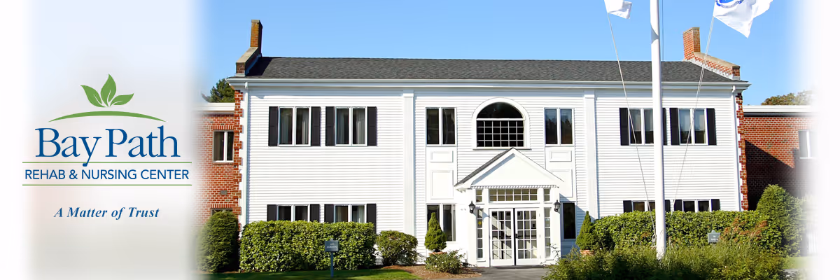 Front exterior view of Bay Path Rehabilitation & Nursing Center building with white siding, multiple windows, a central entrance with a small portico, and surrounding greenery under a clear blue sky.