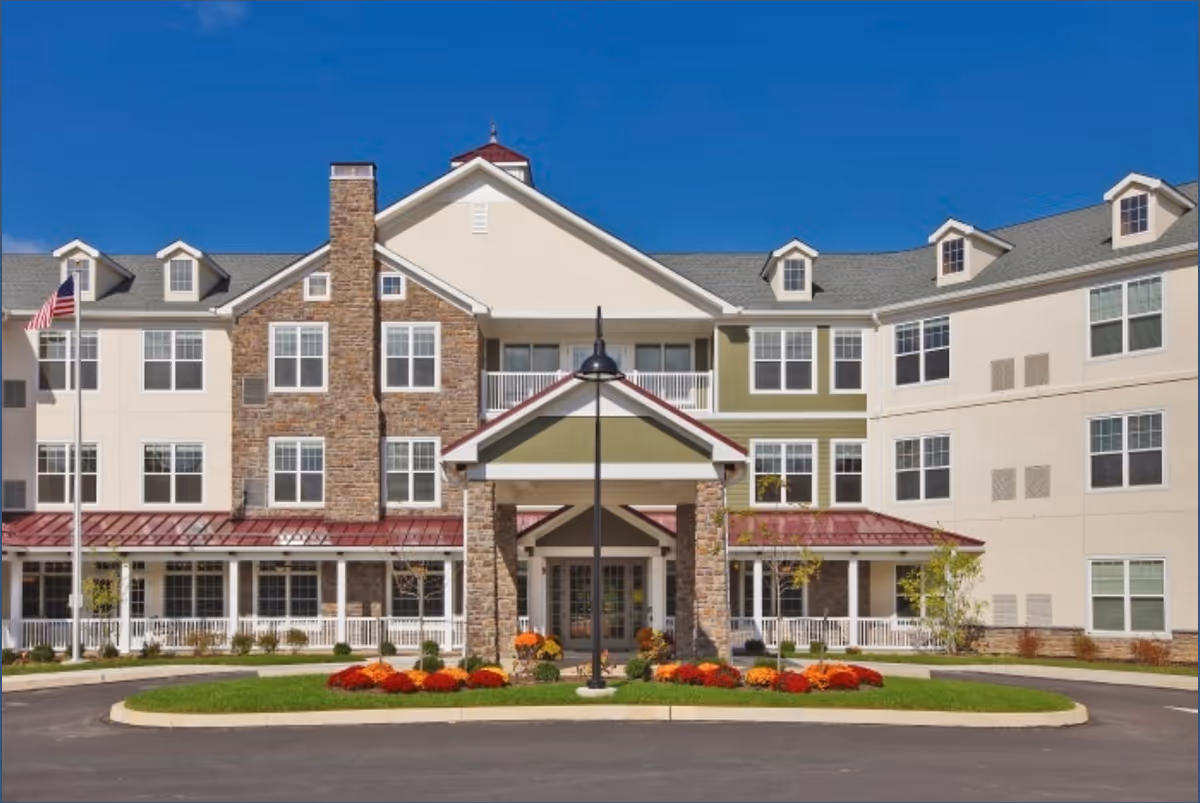 Front exterior view of a multi-story senior living facility building with a covered entrance, stone and beige siding, multiple windows, a flagpole with an American flag, and landscaped flower beds in front under a clear blue sky.