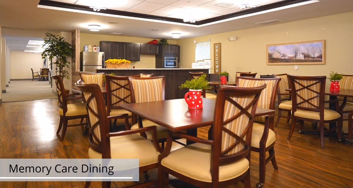 Bright memory-care dining room with wooden tables and chairs, red decorative pitchers, and a kitchen area in the background.