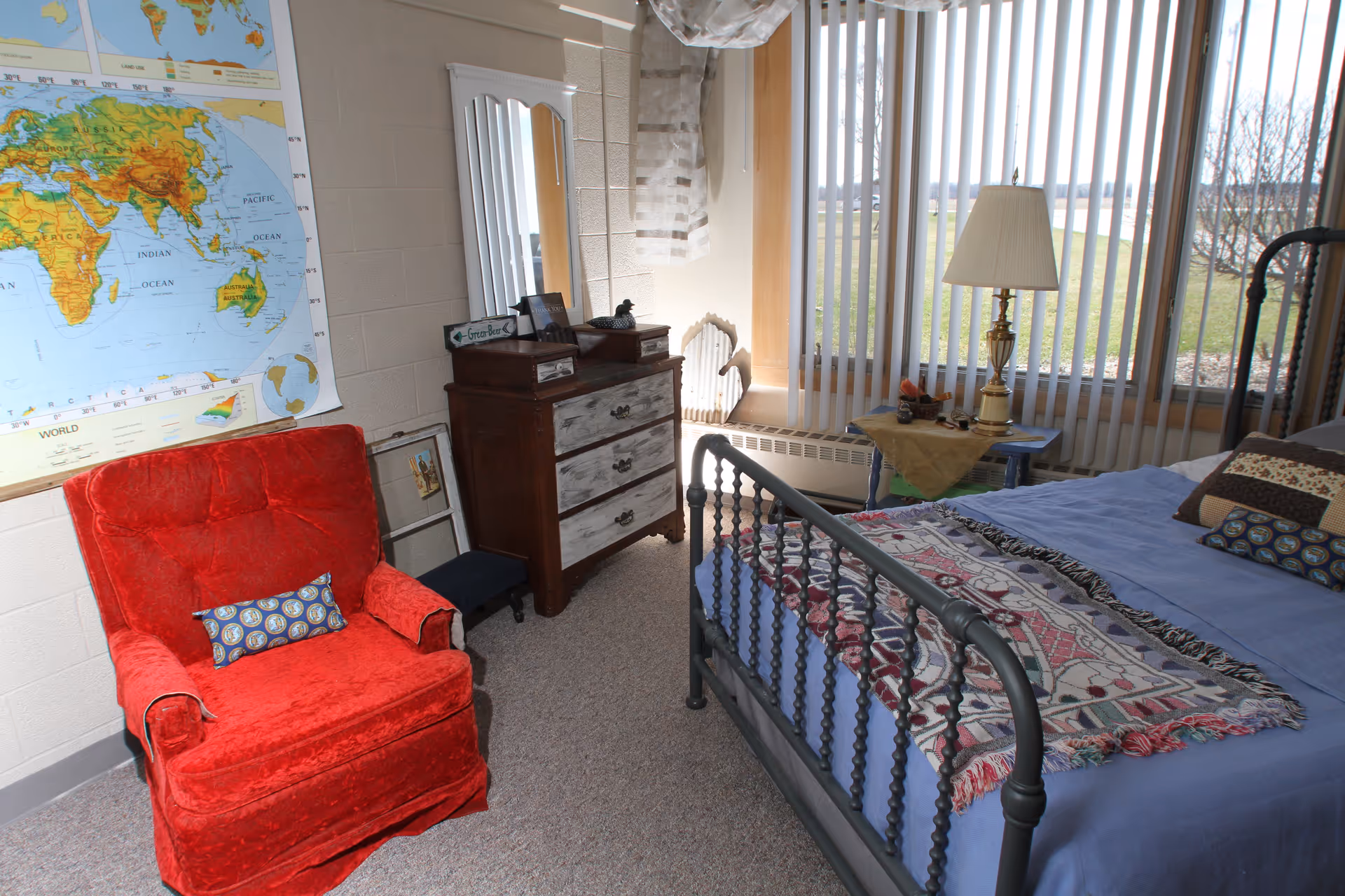 Sunlit bedroom with a metal bed, red armchair, wooden dresser, and a large window with vertical blinds.