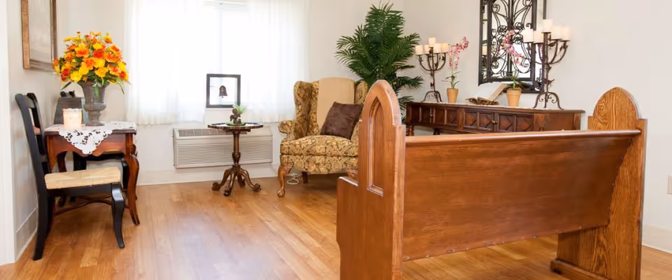 A cozy room with wooden flooring featuring a wooden bench in the foreground, a floral upholstered armchair with a brown pillow, a small round wooden table, and a side table with a vase of yellow and orange flowers. There is a large green plant, a wooden console table with decorative candles, potted plants, and a mirror on the wall. The room is softly lit by natural light coming through a window with white curtains.