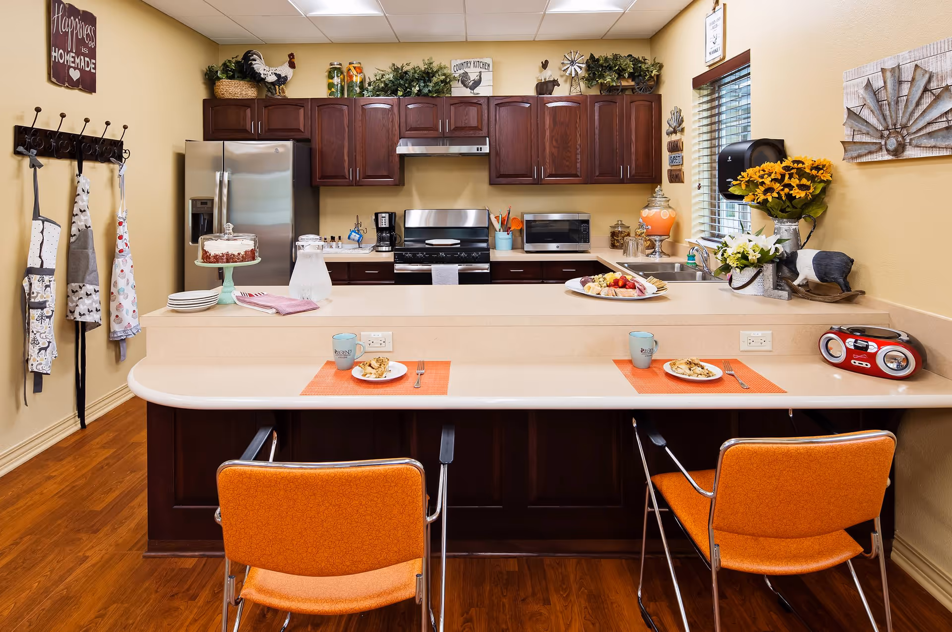 Bright communal kitchen with a long breakfast bar, two orange chairs, stainless steel appliances, and decorative accents.