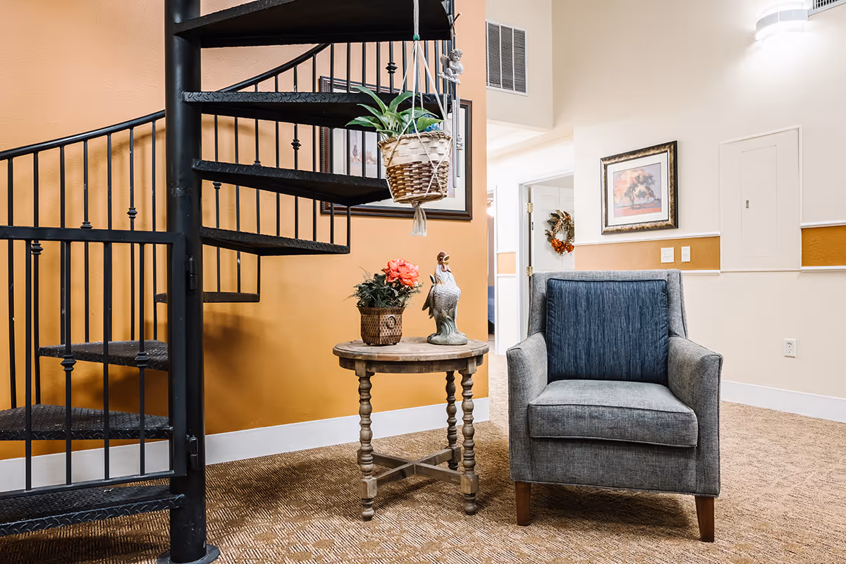 Cozy interior seating area with a gray armchair, round wooden side table holding plants and a decorative statue, and a black spiral staircase against a warm orange wall.