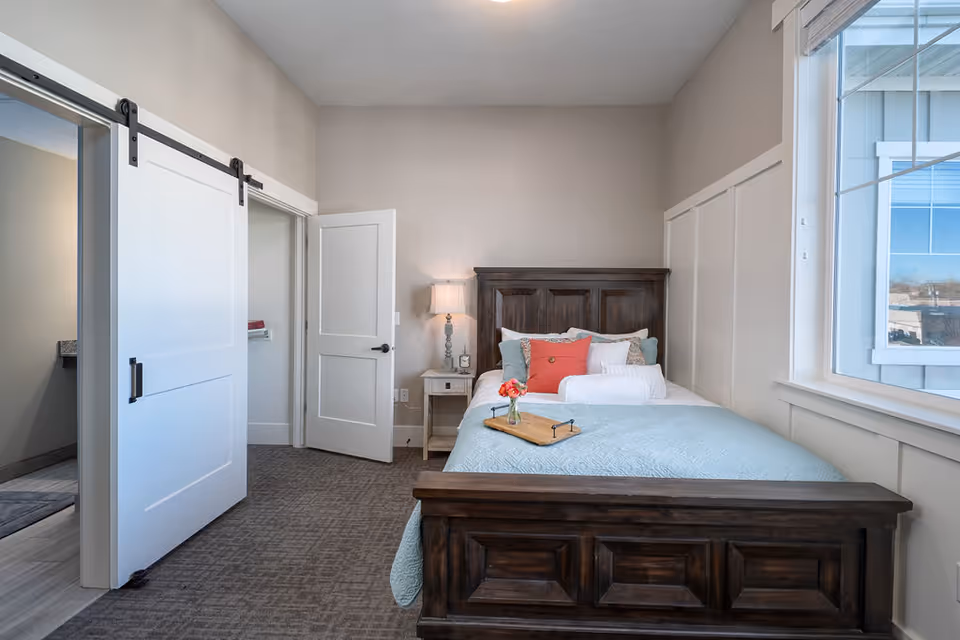 A cozy bedroom in a senior living facility featuring a wooden bed with a light blue quilt, multiple pillows including a coral accent pillow, and a small tray with flowers on the bed. There is a nightstand with a lamp and decorative items next to the bed. The room has beige walls, a large window letting in natural light, and two doors, one of which is a sliding barn door leading to another room.