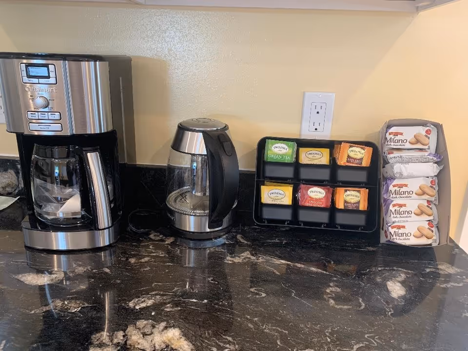 A kitchen countertop with a stainless steel coffee maker, an electric kettle, a black tea organizer containing various Twinings tea bags, and a stack of Milano dark chocolate cookies packages against a beige wall with an electrical outlet.
