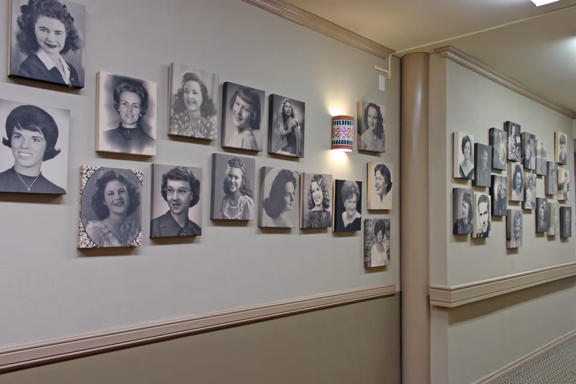 A hallway wall decorated with numerous black and white vintage portrait photographs of various people, mostly women, arranged in a grid pattern. The wall is painted beige with a chair rail and a decorative wall sconce with a colorful pattern is mounted among the photos.