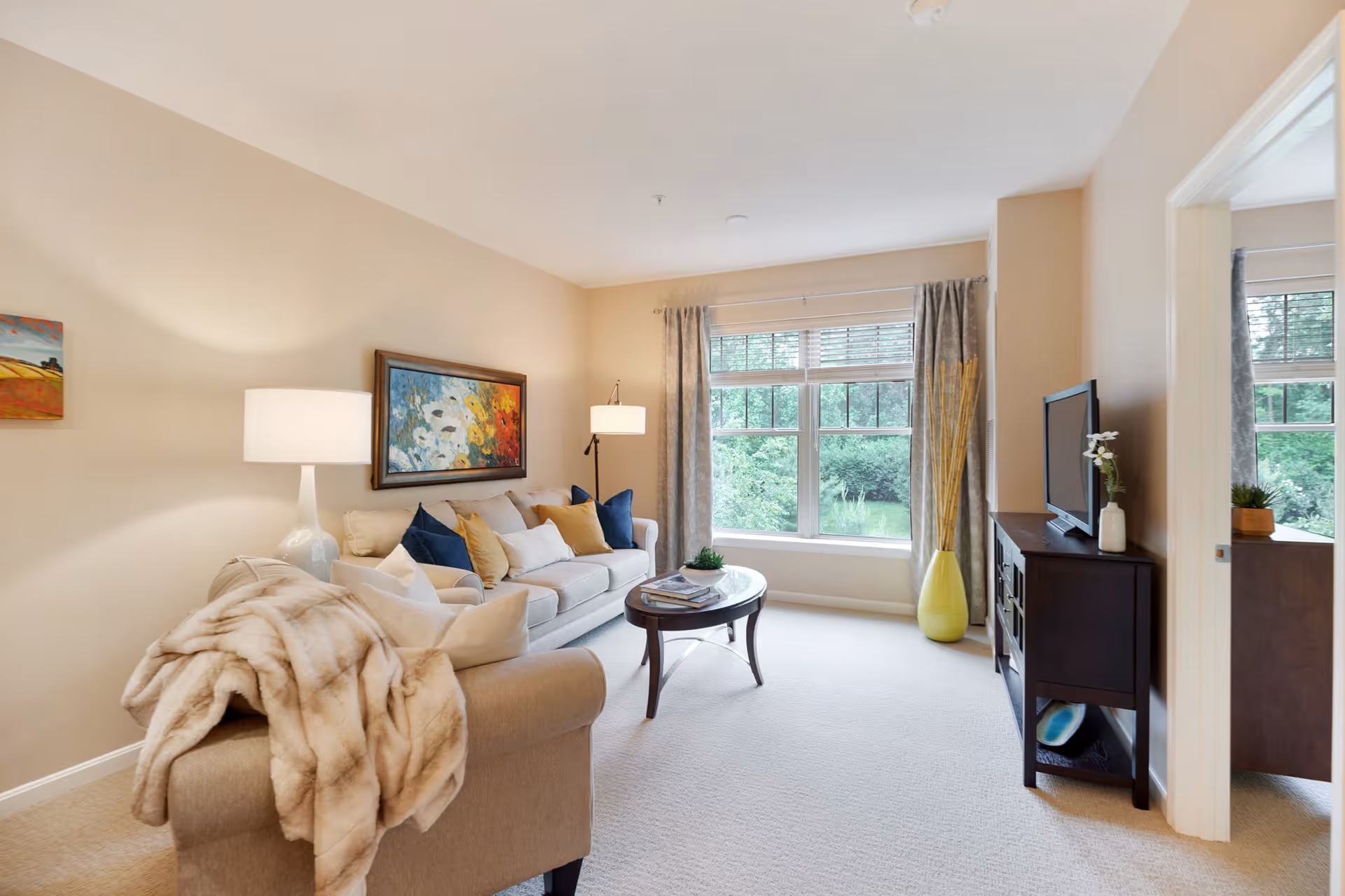 A bright and cozy living room with a beige sofa adorned with multiple cushions and a soft throw blanket. There is a round wooden coffee table in front of the sofa with some magazines and a small plant. A large window with curtains lets in natural light and offers a view of greenery outside. A dark wooden TV stand with a flat-screen TV and decorative items is positioned against the wall. The walls are painted a light beige color, and there are two lamps providing warm lighting. A colorful painting hangs above the sofa.