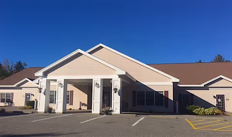 Front exterior view of a single-story beige building with a covered entrance supported by white columns, a brown roof, and several windows. The building is set against a clear blue sky with a parking lot in front.