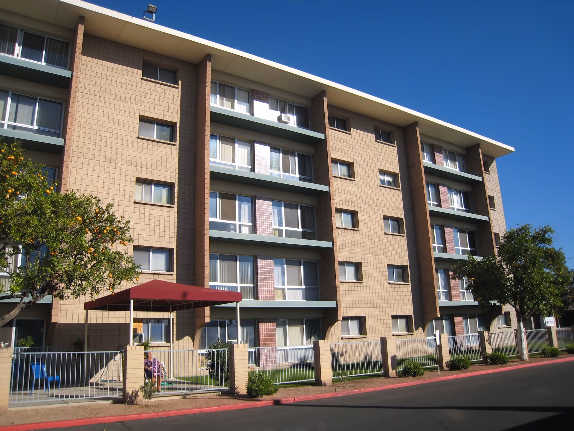 Five-story beige brick senior living building with balconies, a red entrance canopy, fenced front area and trees under a clear blue sky.