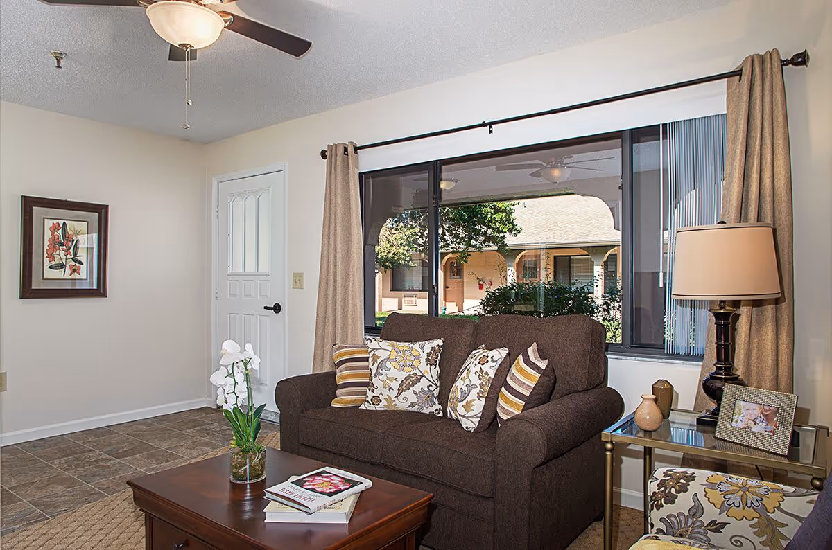 A cozy living room with a brown loveseat adorned with patterned throw pillows, a wooden coffee table with books and a vase of white flowers, a side table with a lamp, decorative vases, and a framed photo. The room has a large window with beige curtains showing an outdoor view of a courtyard with greenery and building arches. A ceiling fan with a light fixture is visible on the ceiling.