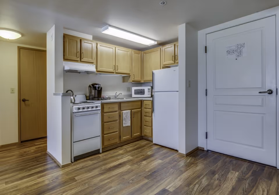A small kitchen area with wooden cabinets, a white stove, a white refrigerator, a microwave, and a coffee maker. The floor is wood laminate, and there is a white door with a floor plan attached to it on the right side of the image.