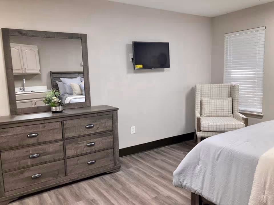 Furnished bedroom with a wooden dresser and mirror, wall-mounted TV, upholstered armchair, and a partial view of the bed.