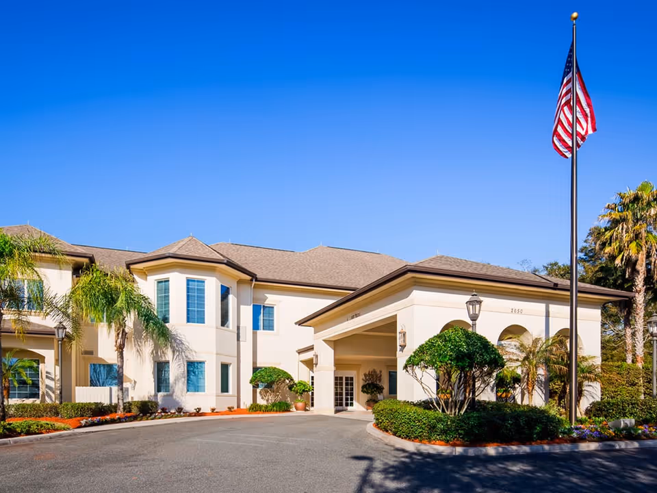 Exterior view of The Windsor Of Ocala building with a clear blue sky, palm trees, manicured bushes, and an American flag on a flagpole in front of the entrance.