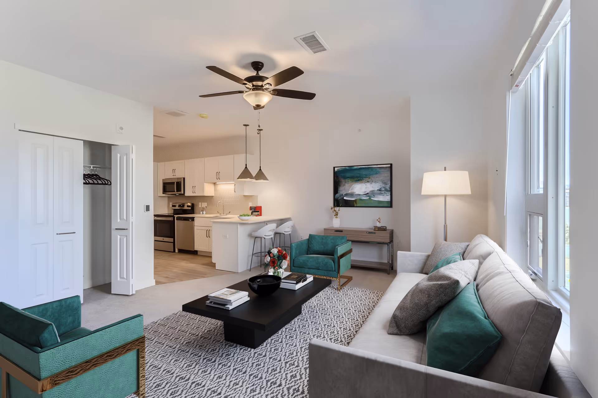 Bright modern living room with a sofa, green armchairs, coffee table and rug, a ceiling fan overhead and an open kitchen with island visible in the background.
