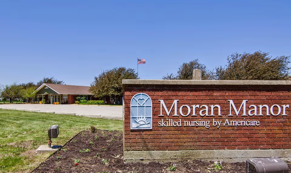 Exterior view of Moran Manor skilled nursing facility with a brick sign in the foreground displaying the facility name and logo, an American flag flying in the background, and a single-story building surrounded by trees and grass under a clear blue sky.