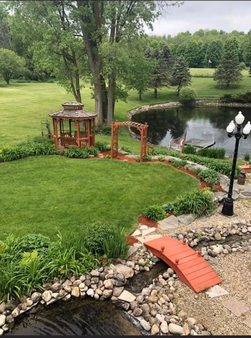 A landscaped outdoor garden with a wooden gazebo, arbor, small red footbridge over a rocky stream, and a pond surrounded by lawn and trees.
