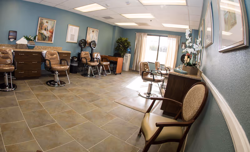 Interior of a salon area in a senior living facility with several salon chairs, hair dryers, a large window with curtains, framed artwork on the walls, and a tiled floor.