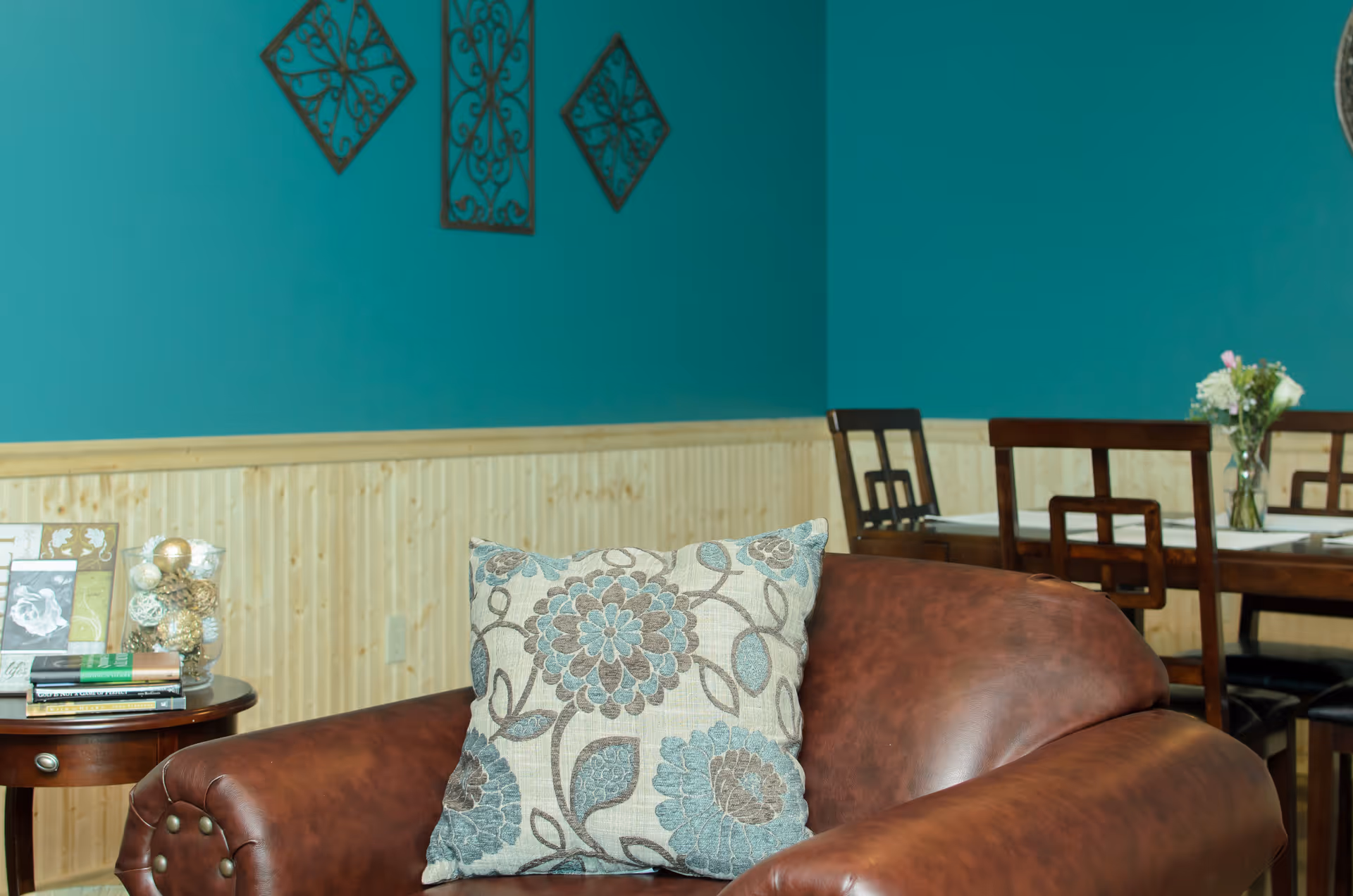 A cozy interior room featuring a brown leather armchair with a floral patterned cushion. Behind the chair is a wooden dining table with chairs and a vase of flowers. The walls are painted teal with wooden wainscoting and decorative metal wall art.