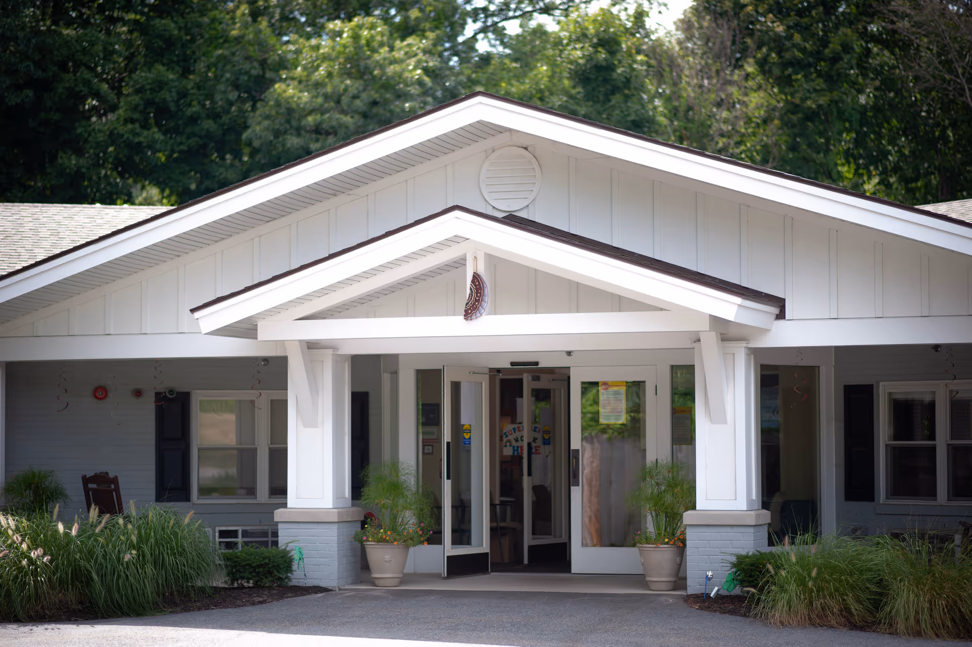 Front entrance of a single-story white building with a covered portico, potted plants, and glass double doors.