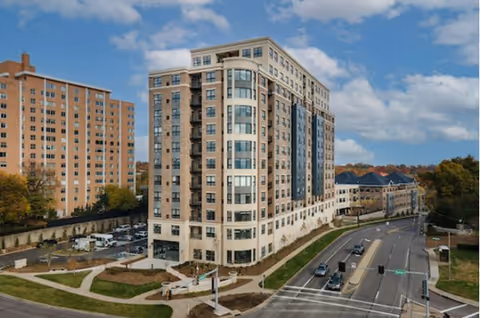 Modern multi-story senior living facility on a curved street corner with blue sky.