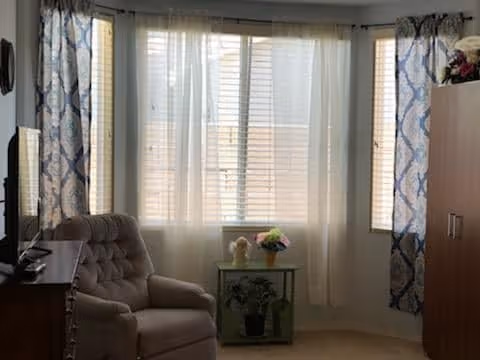 A cozy living room area with a cushioned armchair, a small glass table holding a flower pot and a decorative item, large windows with sheer white curtains and patterned drapes, and a wooden cabinet on the right side.