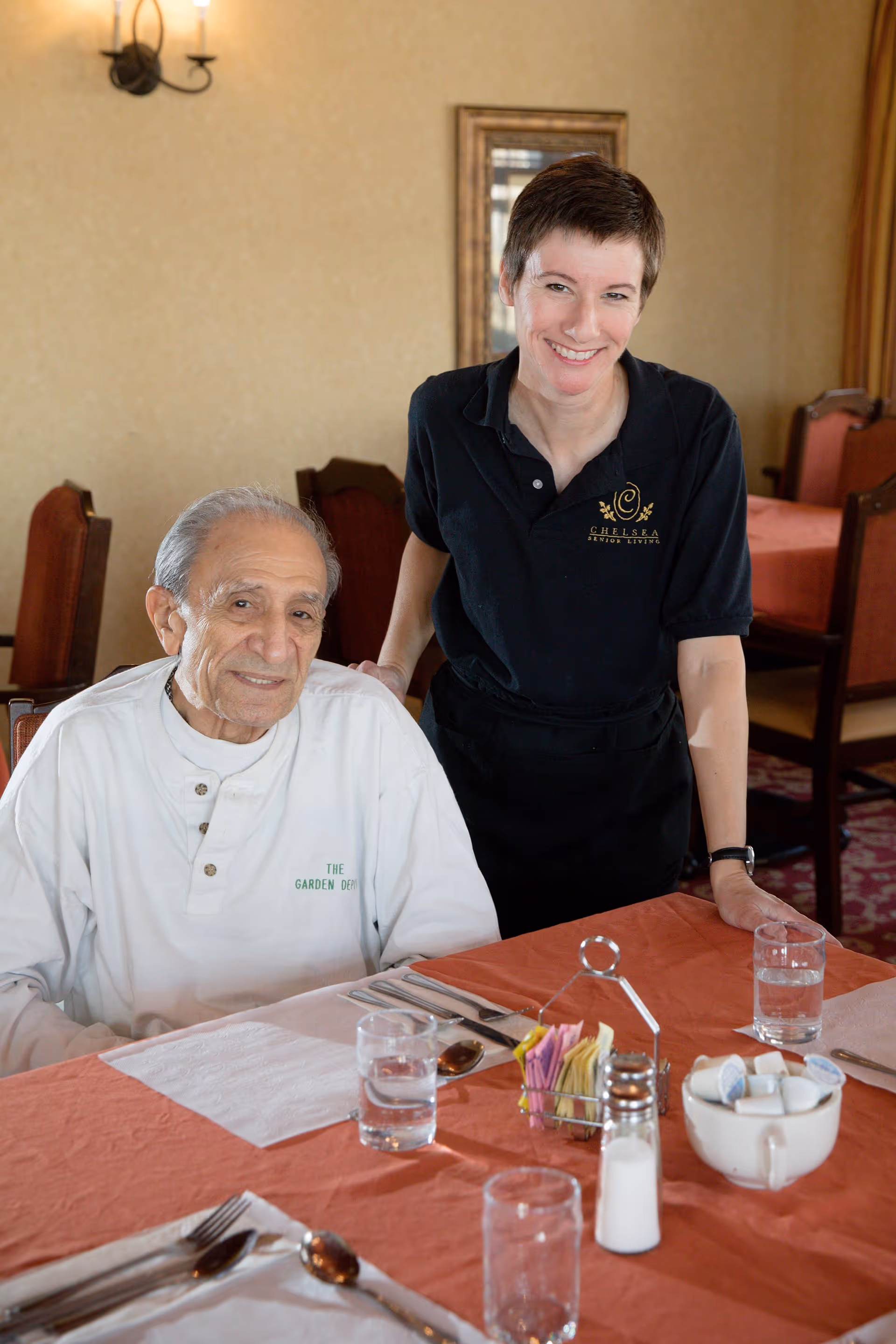 An elderly man sitting at a dining table with a smiling female staff member standing beside him in a dining room. The table is set with glasses of water, utensils, a salt shaker, a container with creamers, and a holder with sugar packets. The staff member is wearing a black polo shirt with a logo that reads 'Chelsea Senior Living'.