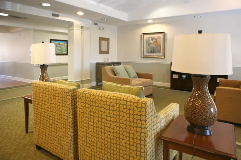 A bright common lounge with patterned armchairs, sofas, side tables and lamps in a senior living facility.