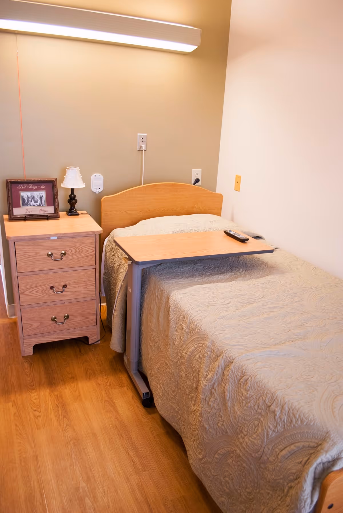 A single bed with a beige quilted bedspread in a room with wooden flooring. Next to the bed is a wooden nightstand with three drawers, a small lamp, and a framed photo. A wooden overbed table with a remote control on it is positioned over the bed. The wall behind the bed has a mounted light fixture and electrical outlets.
