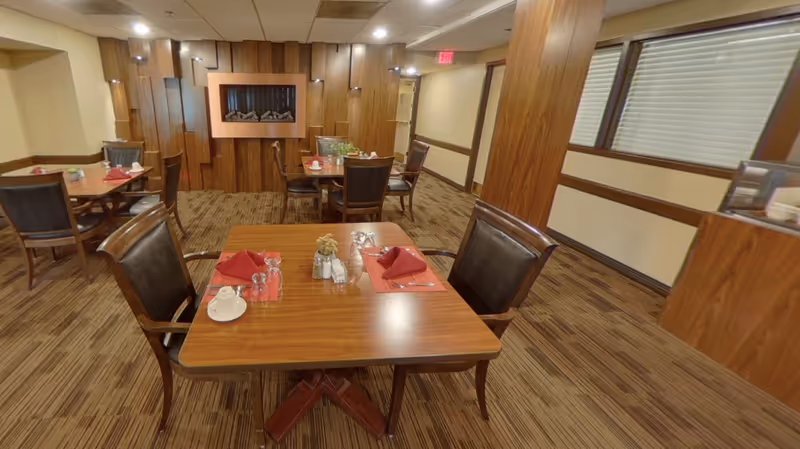 Dining area with wooden tables and chairs set with red napkins and place settings in a senior living facility.