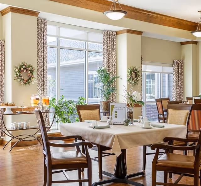 Bright dining room with large windows letting in natural light, decorated with plants and floral wreaths on the walls. Tables are set with white tablecloths, napkins, cups, and a menu card. Wooden chairs surround the tables, and a side table holds beverages and pastries.