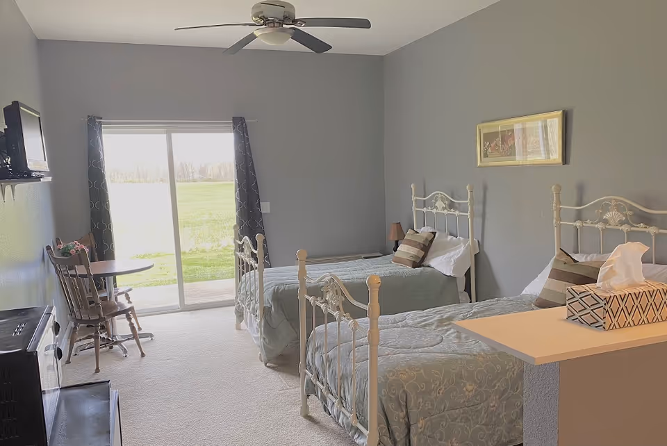 Bright bedroom with two twin iron-frame beds, a sliding glass door, small table and chairs, and a ceiling fan.