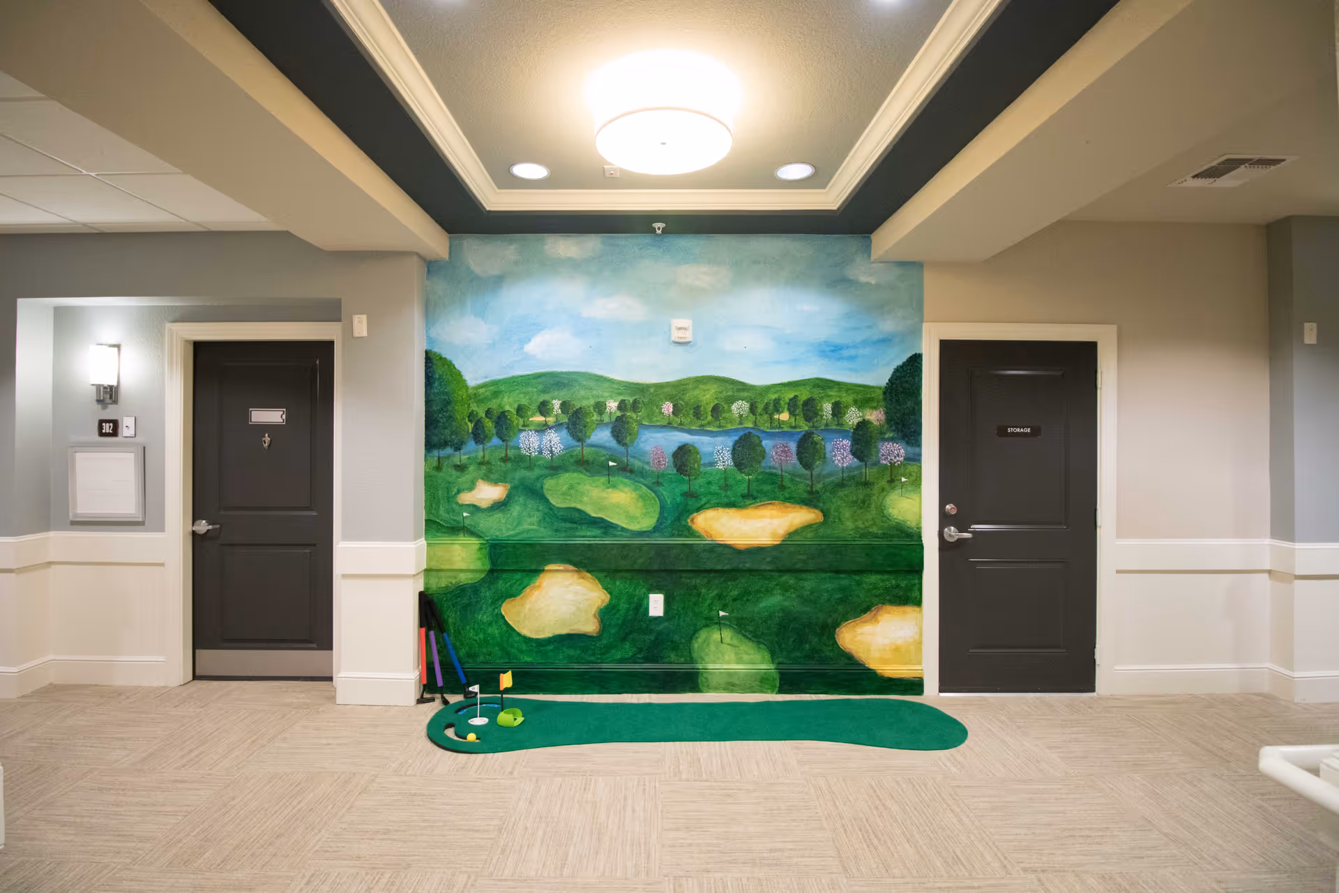 Indoor hallway area with two dark doors on either side of a wall painted with a golf course mural. In front of the mural is a small indoor putting green with golf clubs and balls. The ceiling has a recessed light fixture and the walls are painted in neutral tones.