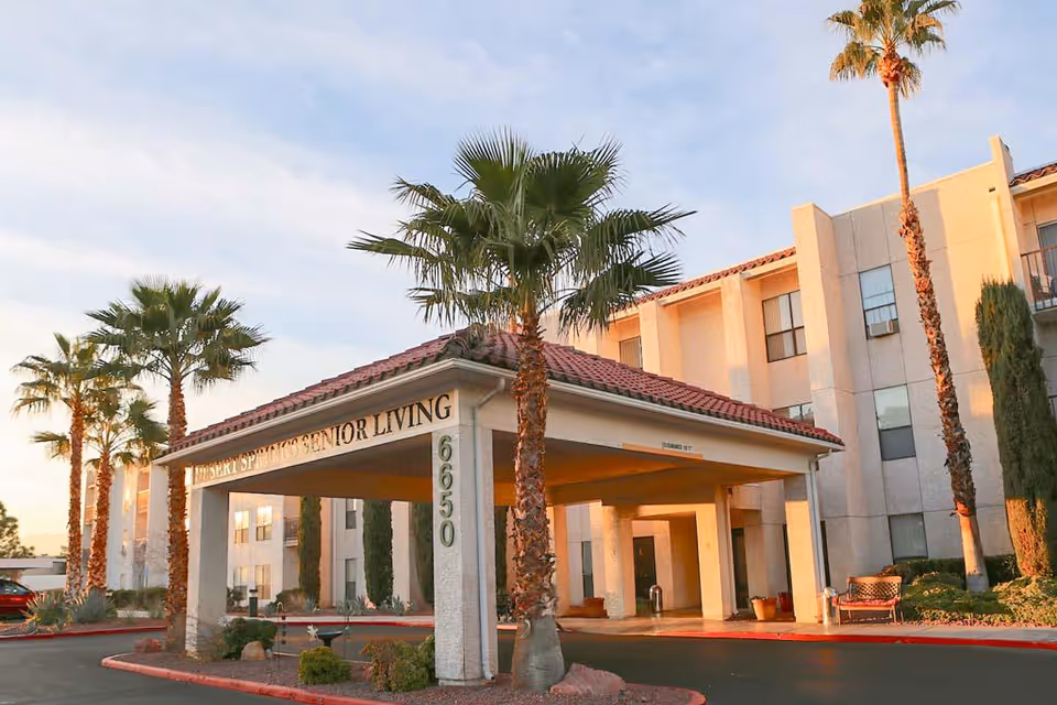 Exterior view of Desert Springs Senior Living facility with a covered entrance, palm trees, and a multi-story building in the background during sunset.