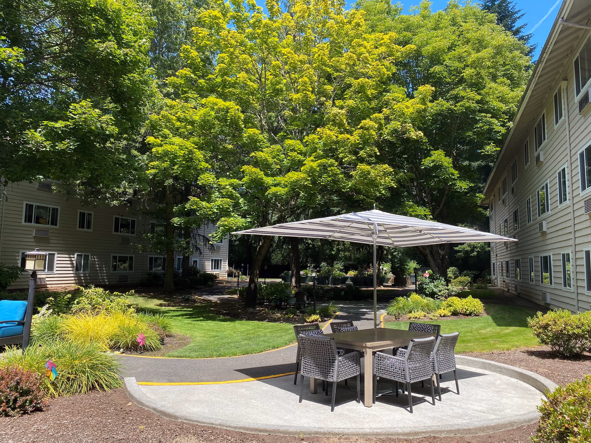 Outdoor seating area with a table and six chairs under a large striped umbrella, surrounded by green trees and bushes, with two beige multi-story buildings on either side under a clear blue sky.