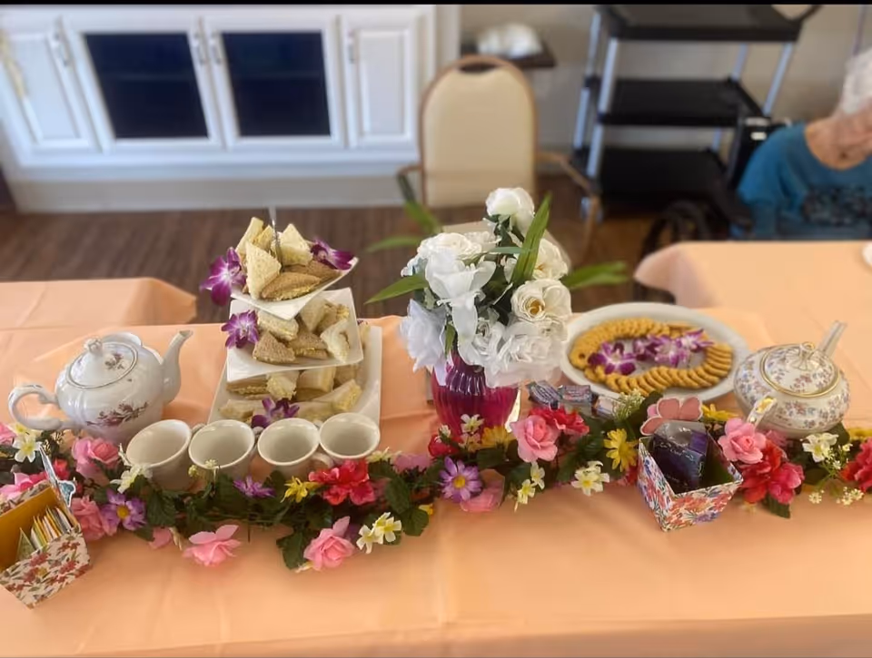 A table set for tea with a three-tiered tray of finger sandwiches decorated with purple flowers, a white teapot with floral design, four white teacups, a vase of white flowers, a plate of crackers with purple flowers, a floral sugar bowl, and a colorful floral garland running along the front of the table. In the background, there is a chair and a person partially visible.