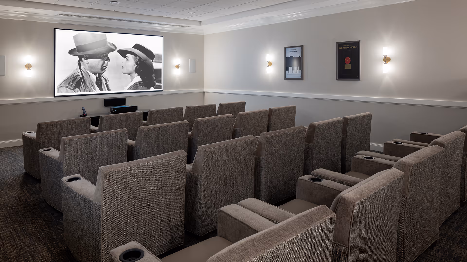 A small movie theater room with three rows of gray upholstered reclining chairs facing a large screen showing a black and white movie scene of a man and woman wearing hats. The walls are light-colored with framed posters and wall sconces providing soft lighting.