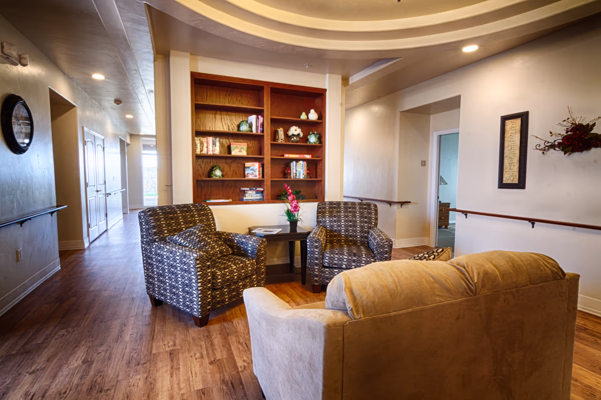 A cozy seating area in a senior living facility with two patterned armchairs and a beige sofa arranged around a small dark wooden table with a flower vase. Behind the seating area is a wooden bookshelf with books and decorative items. The room has wooden flooring, light-colored walls, handrails along the walls, and recessed ceiling lights.
