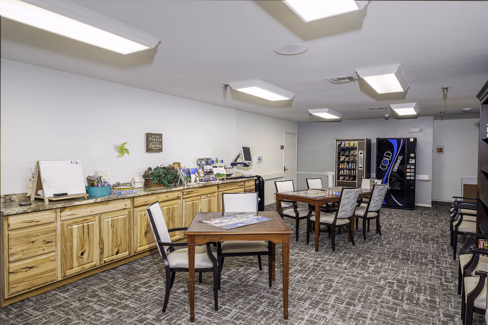 A senior living community activity room with tables and chairs, a long cabinet counter on the left, and vending machines against the back wall.