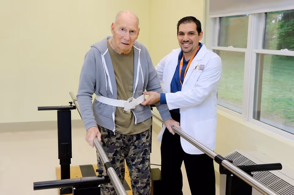 An elderly man wearing a gray hoodie and camouflage pants is using parallel bars for walking support, assisted by a smiling male healthcare professional in a white coat inside a room with large windows.
