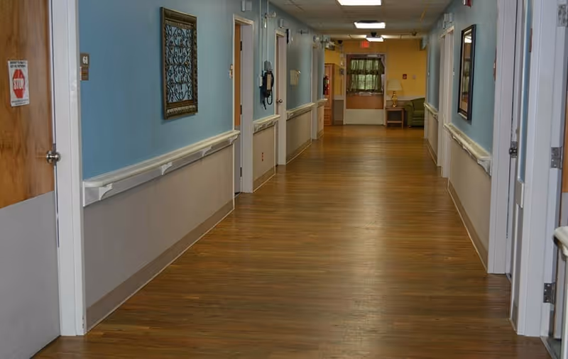 A long indoor hallway with wooden flooring and light blue walls. Several doors line both sides of the hallway, with handrails along the walls. At the far end, there is a small sitting area with a green couch and a lamp on a side table. The ceiling has fluorescent lights and there is a fire extinguisher mounted on the right wall near the end of the hallway.
