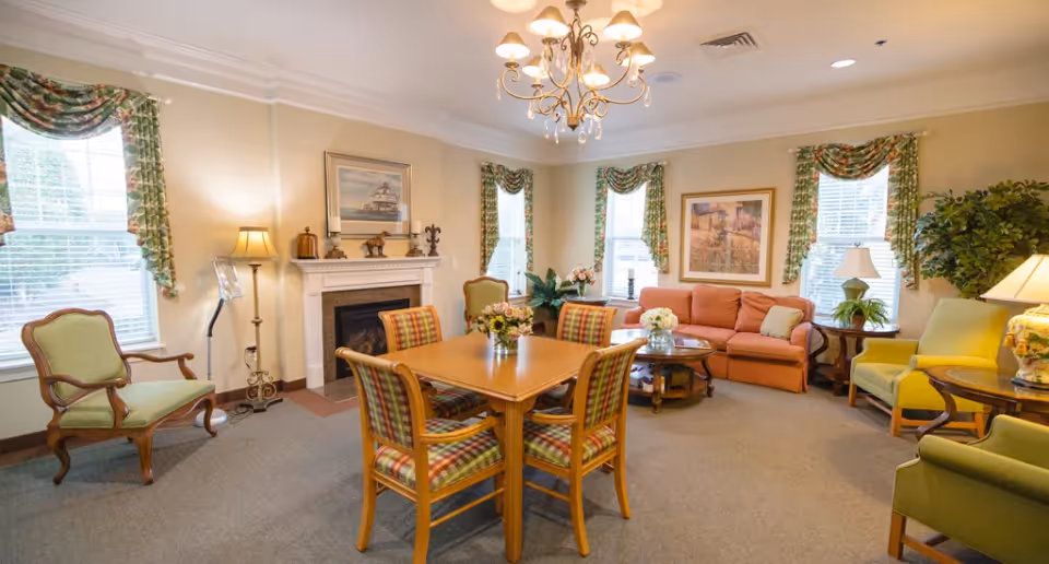 A cozy living room with a wooden table and four plaid cushioned chairs in the center. The room features a fireplace with decorative items on the mantel, a peach-colored sofa, green armchairs, a round coffee table with flowers, and several windows with floral curtains allowing natural light to enter. The walls are adorned with framed paintings, and a chandelier hangs from the ceiling.