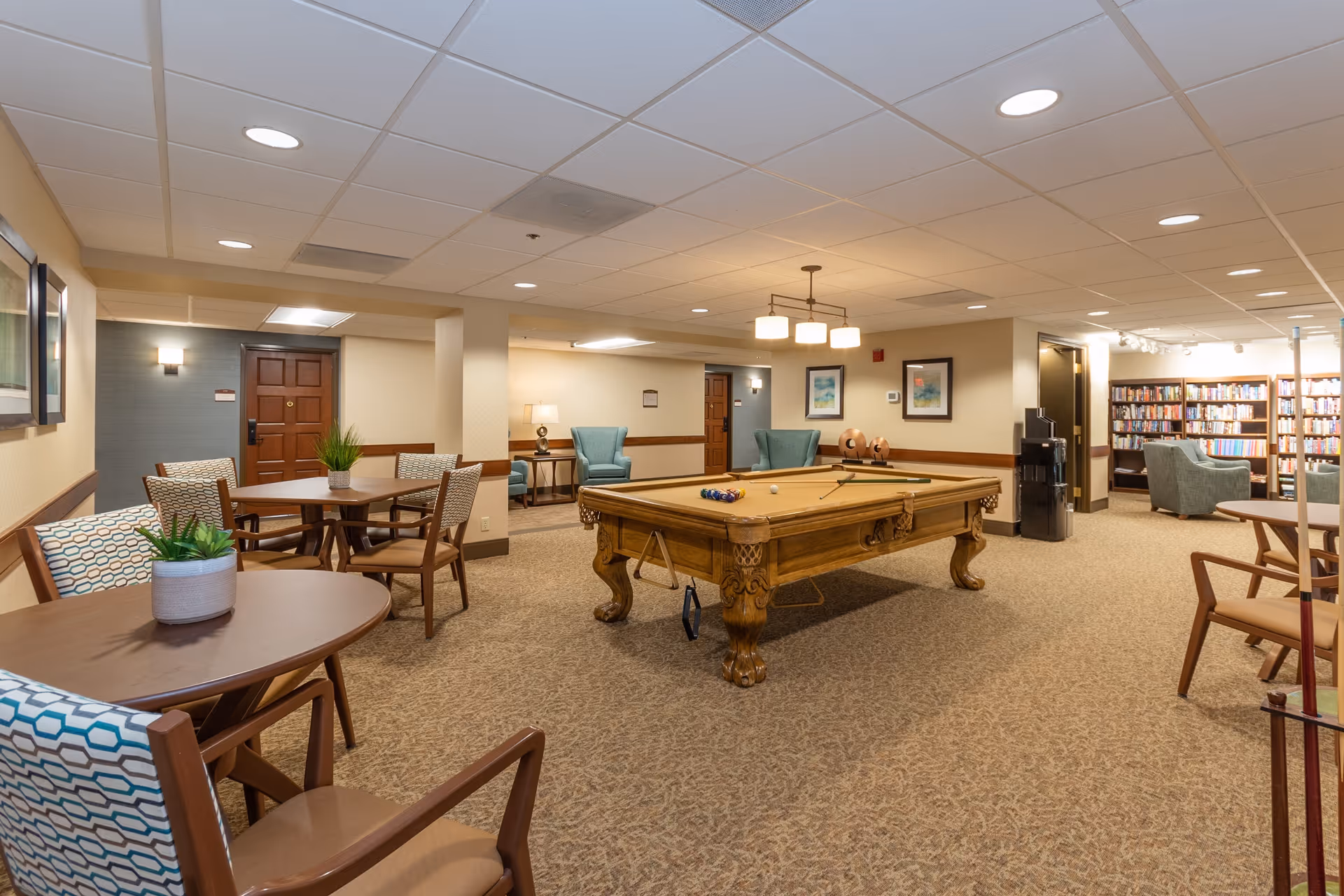 Carpeted communal activity room with a pool table surrounded by chairs, tables, and bookshelves.