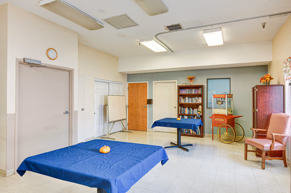 A community room with two tables covered in blue tablecloths, each with a small pumpkin on top. There is a popcorn machine on a red cart, a bookshelf filled with books, a whiteboard on a stand, and a pink armchair near a window with floral curtains. The walls are painted beige and light blue, and there are several doors and a clock on the wall.