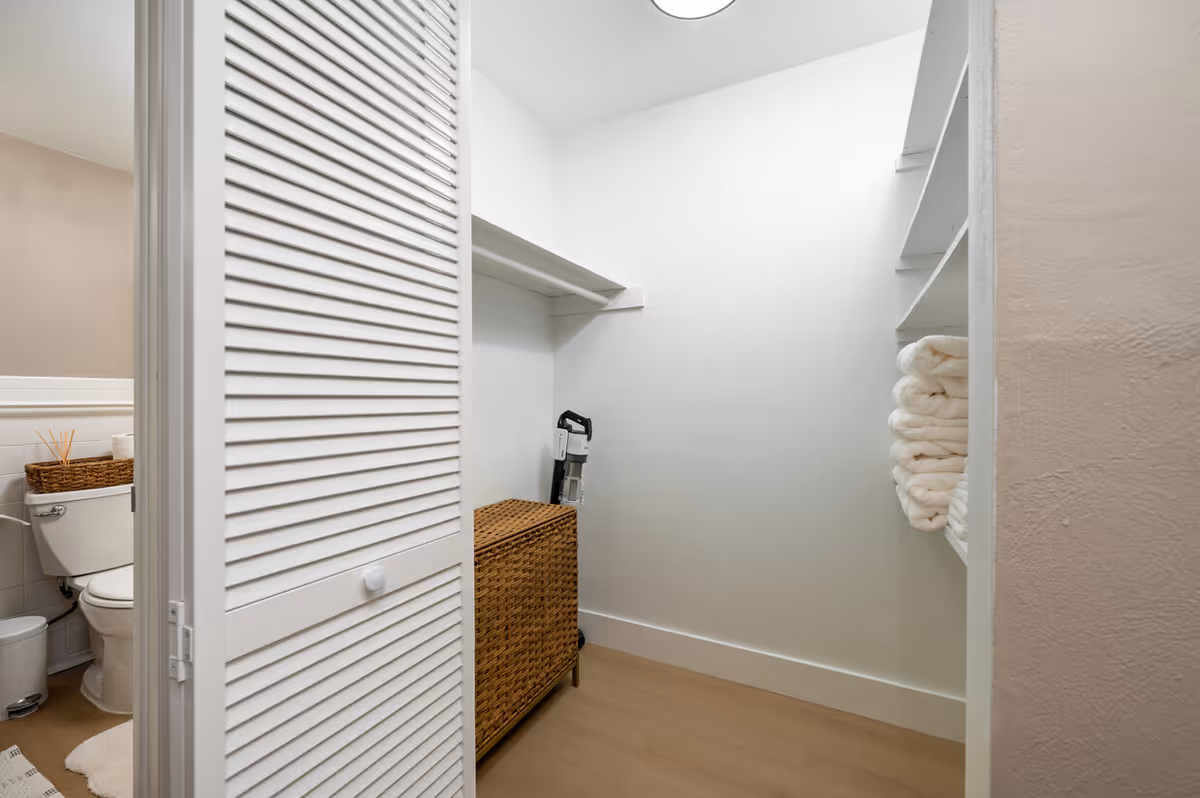 Open linen closet with a white louvered door, folded towels on shelves, a wicker hamper and a partial view of a bathroom toilet.