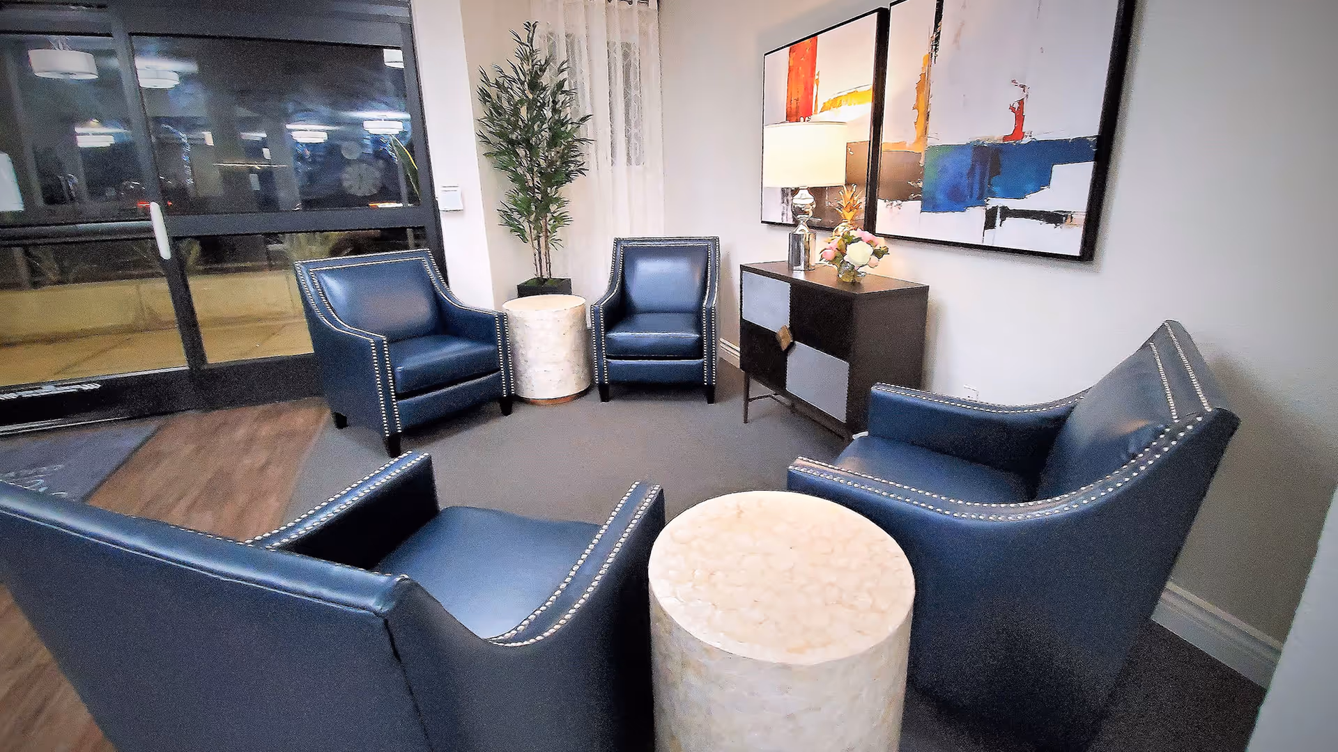 A cozy seating area with four blue leather armchairs arranged around two round white marble side tables. There is a small cabinet with a lamp and a flower arrangement on top, and two abstract paintings hanging on the wall. A tall green plant is placed near a glass door entrance.